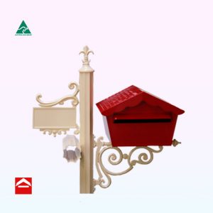 Chalet style letterbox rear open on a scroll bracket to the right of a central post. Atop the post is a fleur de lys. To the left is a number plate and newspaper holder.